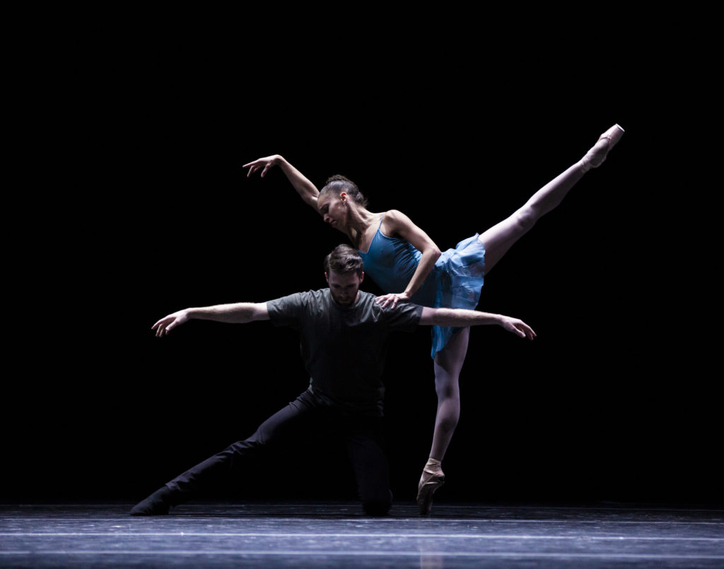 On a darkened stage, Chyrstyn Fentroy balances in first arabesque, her front arm raised to her head in a gesture reminiscent of Swan Lake. She rests her other arm on Roddy Dobble, who hovers in a side lunge, facing forward, with his arms floated to the side, gaze downturned.