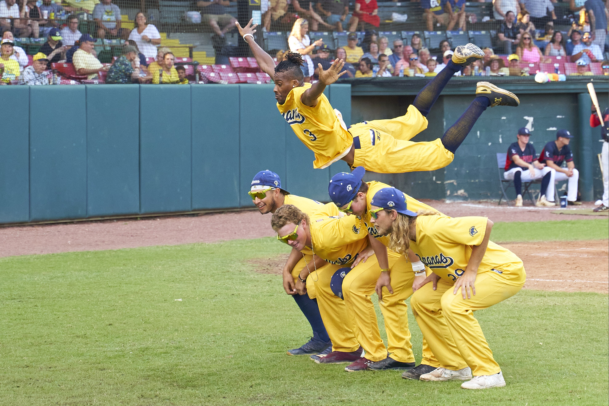 Five men in matching bright yellow baseball uniforms pose together on the field. Four of them crouch with their hands on their knees, while a fifth, Maceo Harrison, flies parallel to the ground over their backs, knees bent behind him and arms splayed out at his sides.