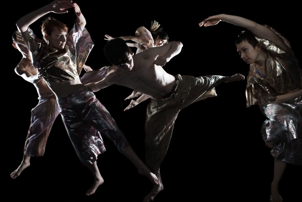 Five dancers in baggy, iridescent costumes fly, shift, and lean to the left of the space framed by the photo, the floor and backdrop a black void.