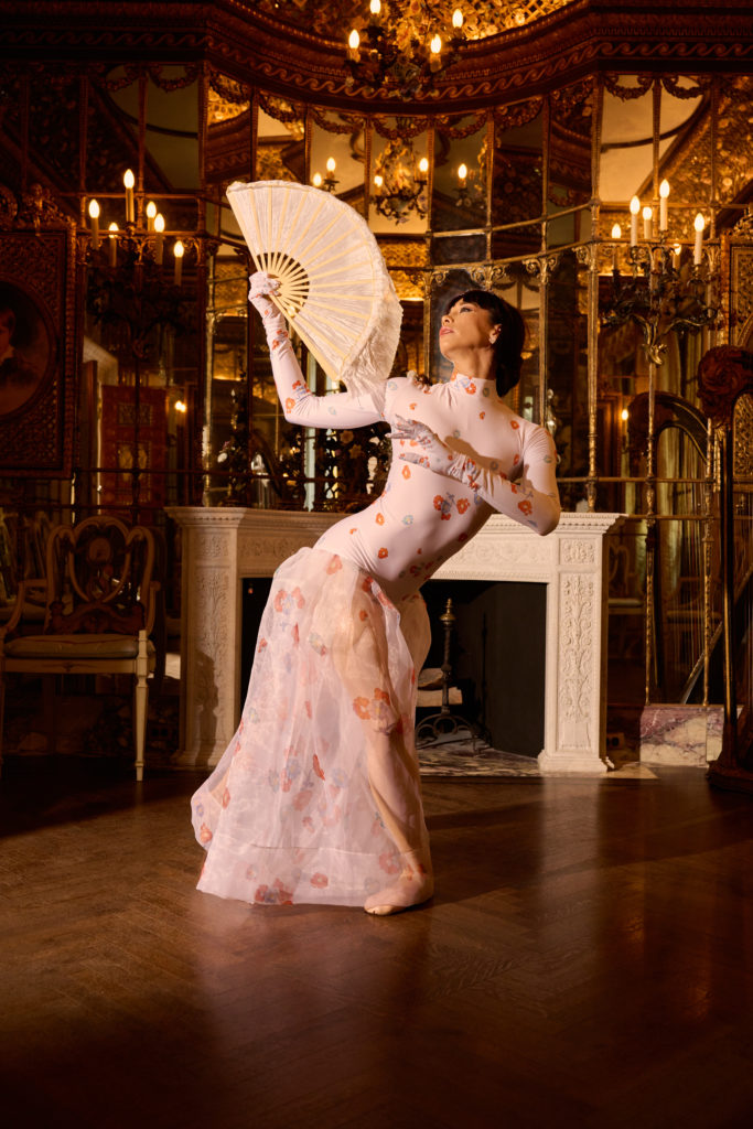 Georgina Pazcoguin poses elegantly in a fourth position lunge while holding open a delicate, lacy folding fan. She is in an ornate, gilded room full of candelabras and mirrors. 