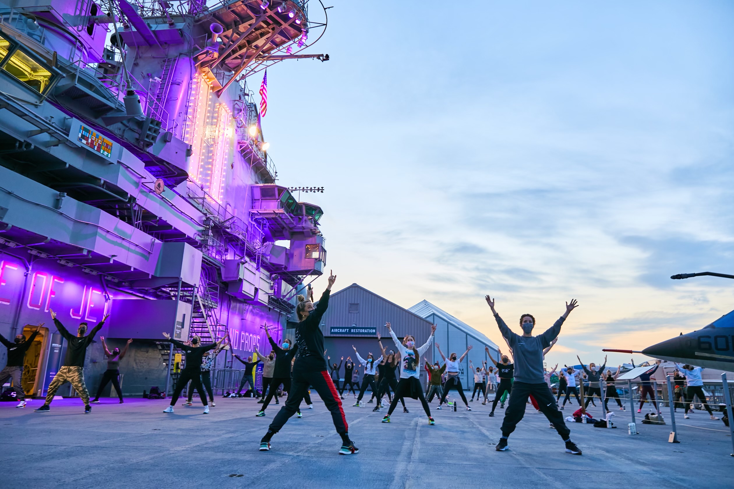 A group of people in workout clothes and face masks stand with their legs spread, arms stretched overhead in a "V," on the deck of a massive ship. The sky's colors change with the advent of dawn or dusk.