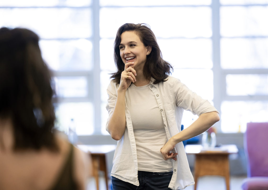 Lauren Lovette brings a hand to her chin as she smiles. Her brown hair is curling loosely to her shoulders. She wears a pale tank top and white cardigan. Bright light streams through windows behind her; in the foreground, the torso of a blurry dancer.