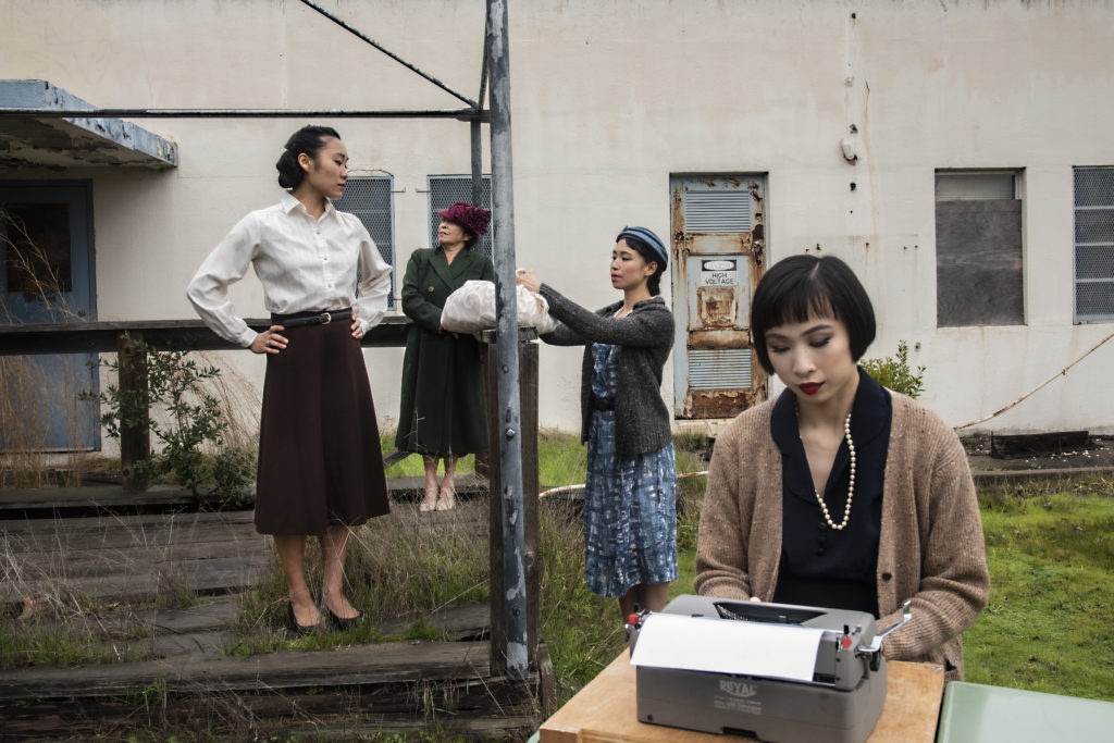 An Asian woman with a chic black bob in 1940s period-wear sits at a typewriter. Three other women, similarly dressed, pose in the overgrown outline of what used to be a building behind her.