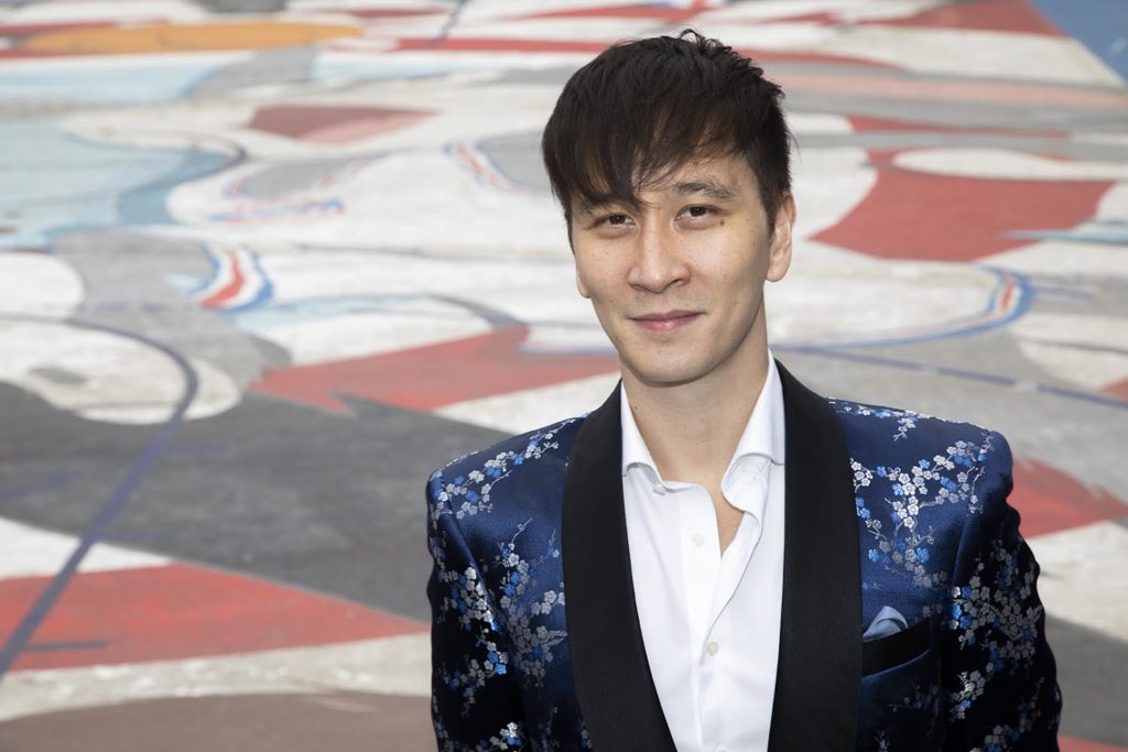 Phil Chan gives a closed mouth smile to the camera. He wears a dark blue sequined suit jacket over an open-collared white button-down. Colorful abstract patterns are visible on the ground behind him.