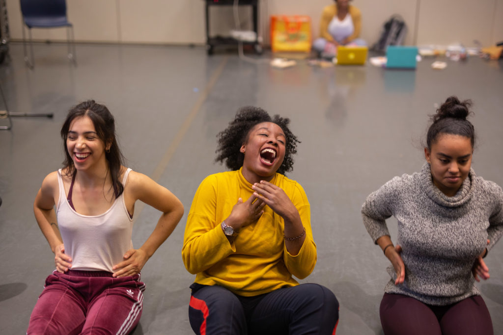 Three dancers smile and laugh as they sit on the floor of a studio. They sit side by side, legs outstretched in front of them, hands coming to their rib cages.