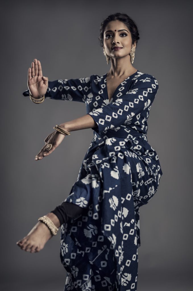 Narayan poses in a navy dress with white print. Her hands are flexed in opposite ways and her right foot is pointed.