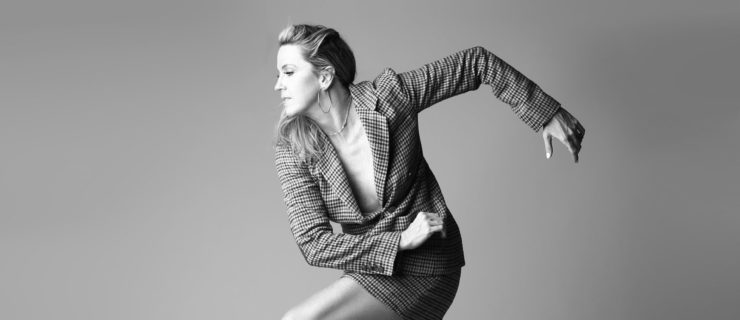 In a black and white image, Kristin Sudeikis is shown in profile, balancing with one foot lightly raised off the floor as her arms wrap into a geometric shape behind her. She wears a long-sleeved, short dress with a plunging neckline and chunky heeled boots.