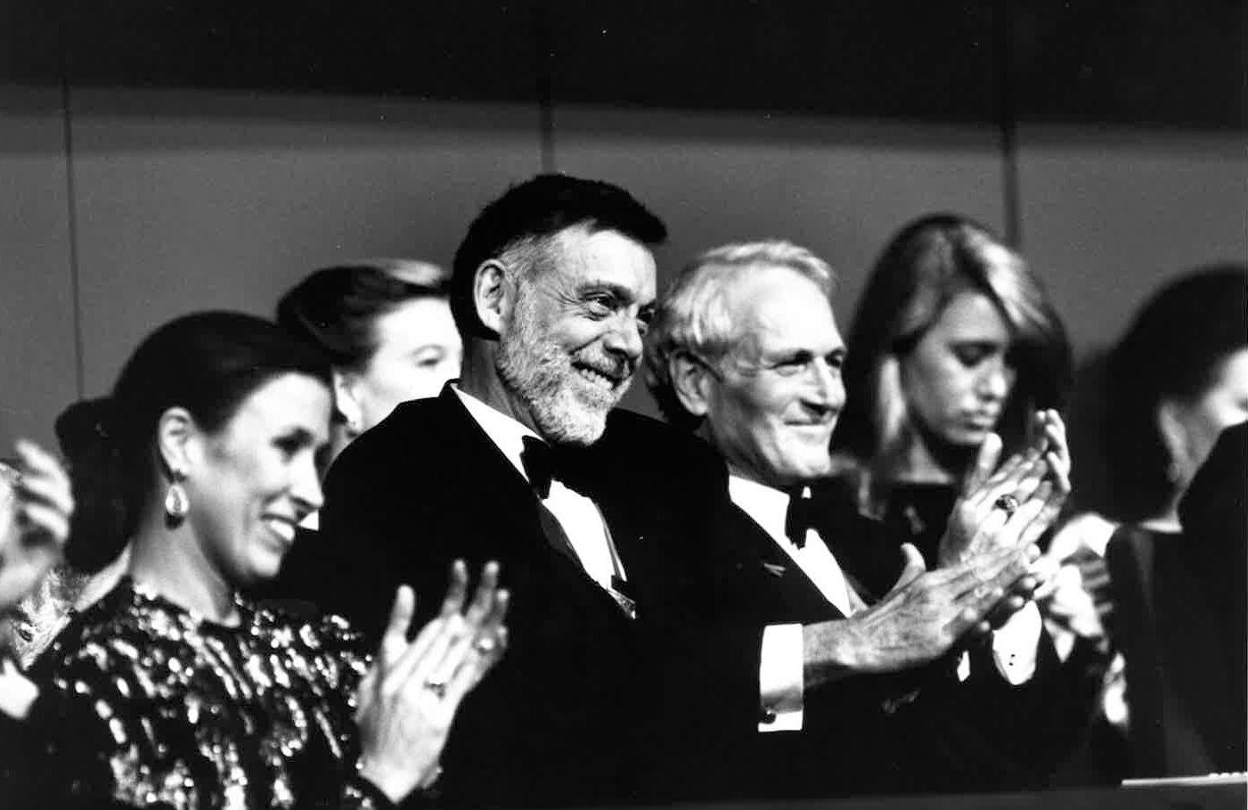 In a black and white archival photo, Paul Taylor sits in a tuxedo in an audience, smiling broadly as he applauds.