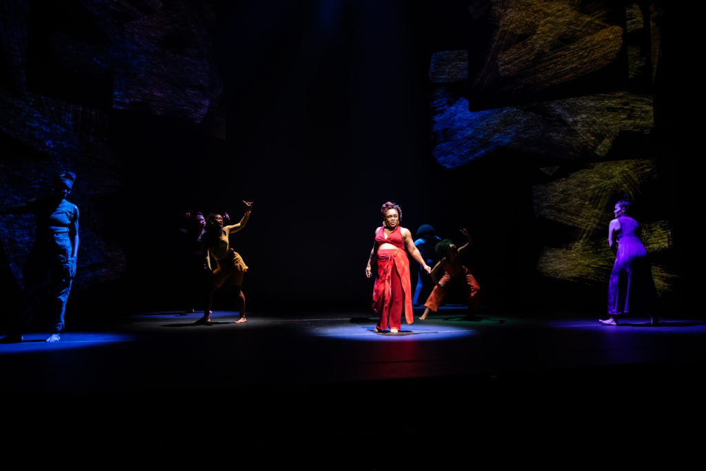 Kenita R. Miller, in a red costume, in spotlit at center stage. Six other performers are lit behind and around her in dark tones.