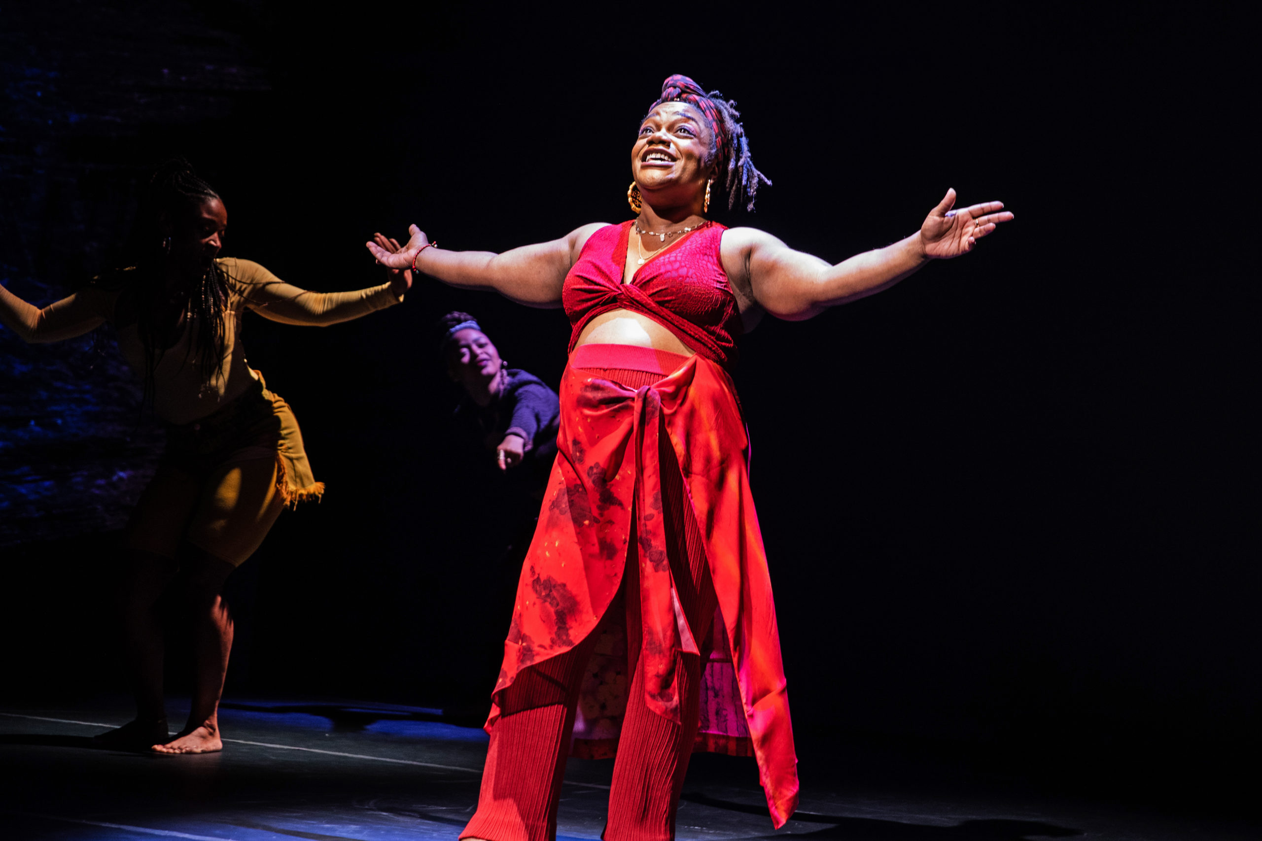 Kenita R. Miller, a Black pregnant Broadway star, extends her arms wide and beams onstage. She is wearing a red crop top and pants, and her pregnant belly is visible.