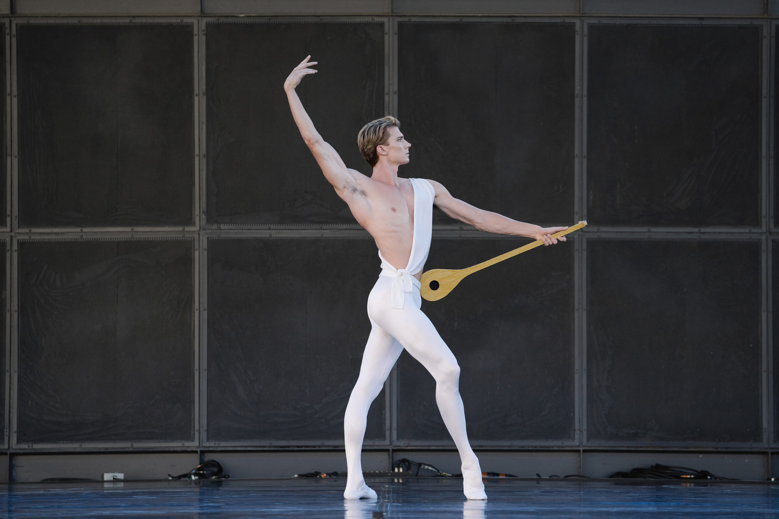 Brendan Saye, costumed in white, poses alone onstage, a prop that evokes a lyre resting against one hip. He stands in fourth with his front foot in forced arch, opposite arm in an extended fifth position behind his head. He looks regally towards the wings, face in profile.
