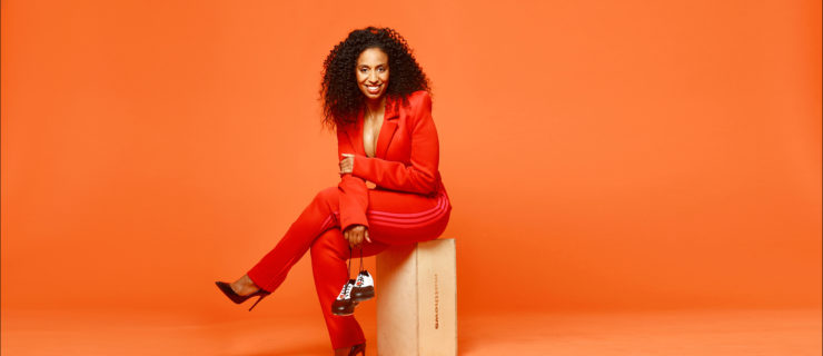 Chloé Arnold is a Black woman with long, dark curly hair. She wears a red blazer and pants and black high heels, and sits facing the camera and smiling, holding black-and-white tap shoes.