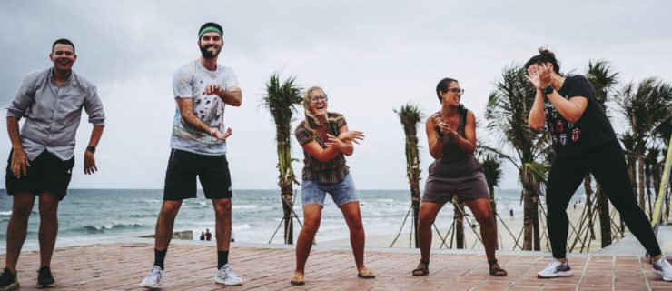 Ethan Levy stands in a line with four other people, all wearing comfortable street clothes as they laugh and imitate each other's gestures. A beach is visible behind them.