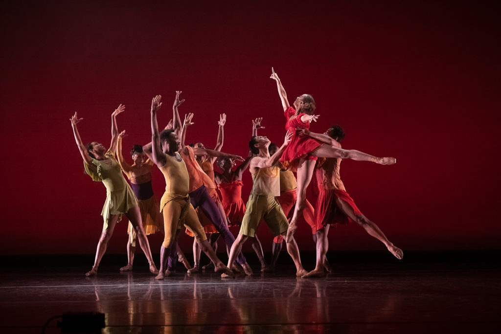 A female dancer is lifted in arabesque on an upstage diagonal, pointing up. One dancer is in the process of tossing her to another. A cluster of dancers arrayed around them turn their gazes up, arms rising as they lung back, away from the lifted dancer.