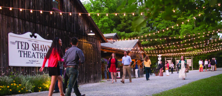 Spectators stroll along a path outside a dark wood barn with a large sign declaring it to be "The Ted Shawn Theatre, 1942." Fairy lights are strung up overhead, while treetops surround the area.