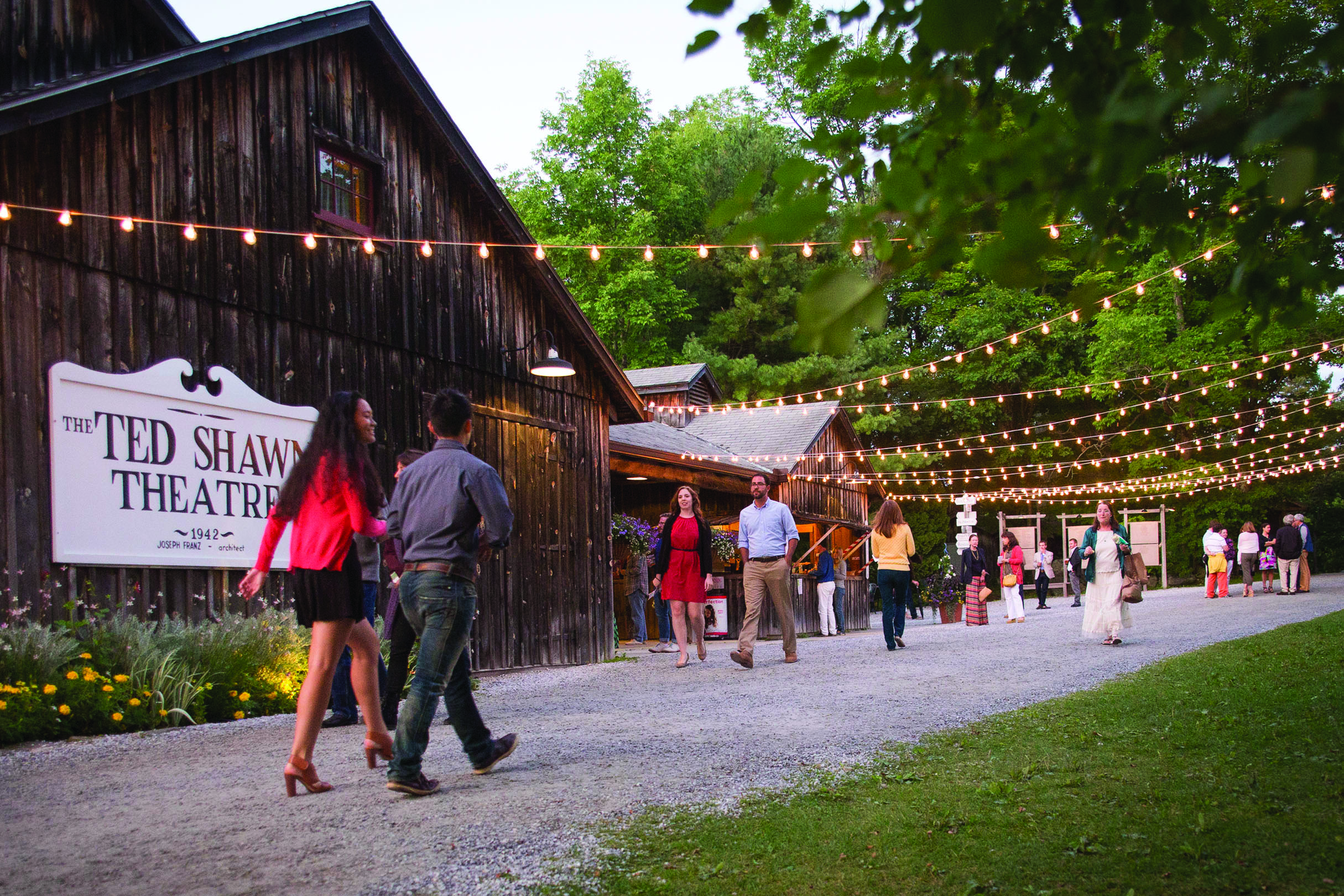 Spectators stroll along a path outside a dark wood barn with a large sign declaring it to be "The Ted Shawn Theatre, 1942." Fairy lights are strung up overhead, while treetops surround the area.