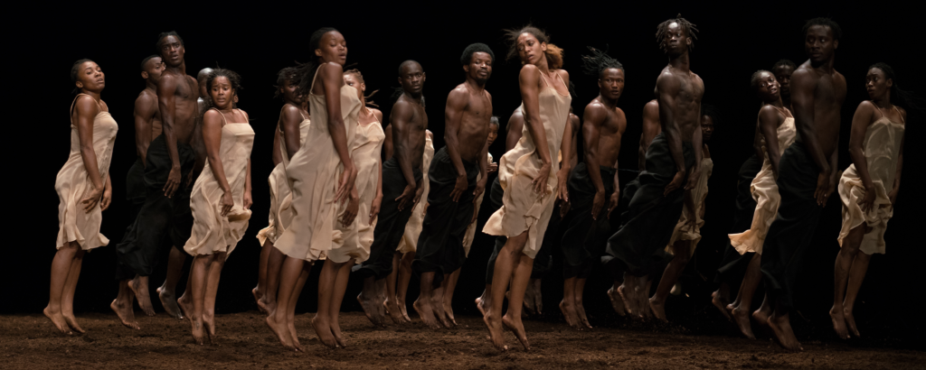 Roughly two dozen Black dancers jump straight up in profile, palms pressed to the tops of their thighs as they turn their heads over their right shoulders. The floor beneath them is loosely packed brown soil. The women wear light beige shifts, the men black trousers and bare chests.
