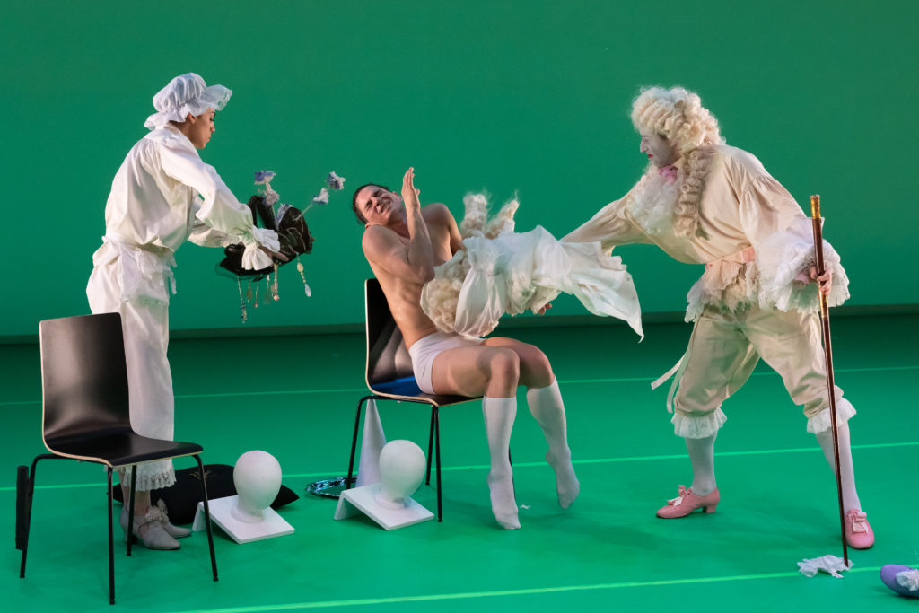 Two male and one female dancer in Baroque costumes and wigs play with props that look like white fabric and a wooden cane. The center dancer is naked except for socks and shorts.