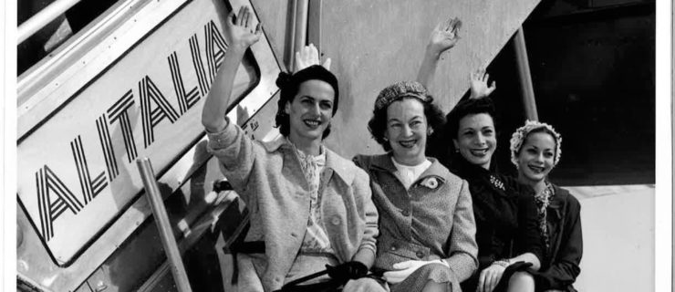 In a black and white archival image, four women dressed in dresses, hats, coats, and heels appropriate to the '50s smile and wave. They are seated, legs crossed, in a line on the supports of a metal stairway, which is rolled up beside an airplane.