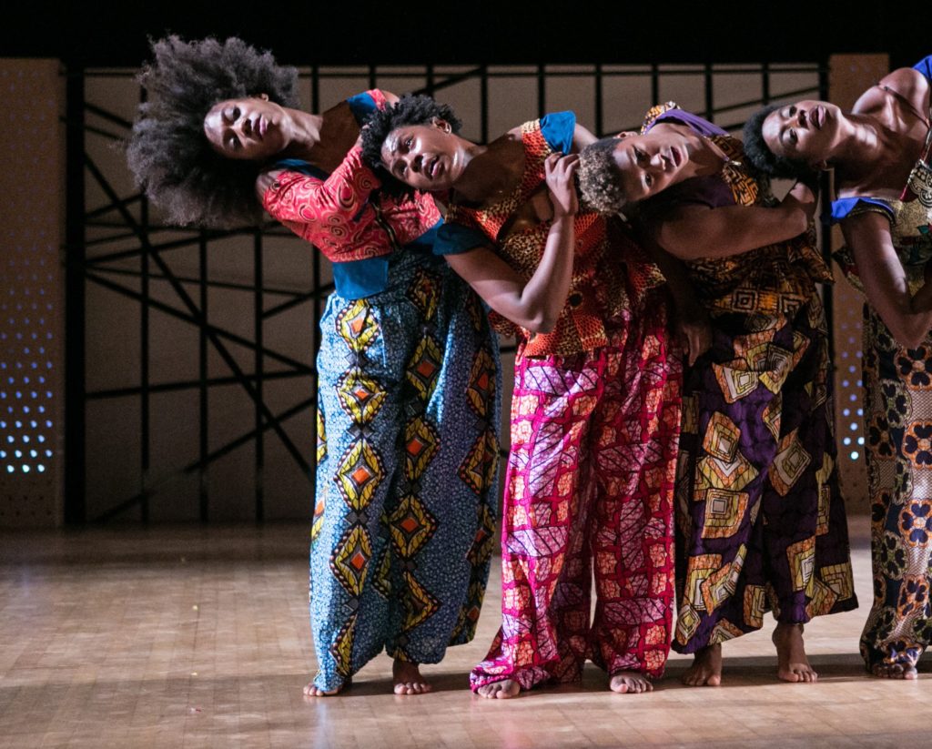 Four Black women stand shoulder-to-shoulder, all leaning their torsos to their right as their arms tuck against their chests, resisting gravity. All four wear different brightly colored and patterned loose trousers and tops; their hair is left natural.