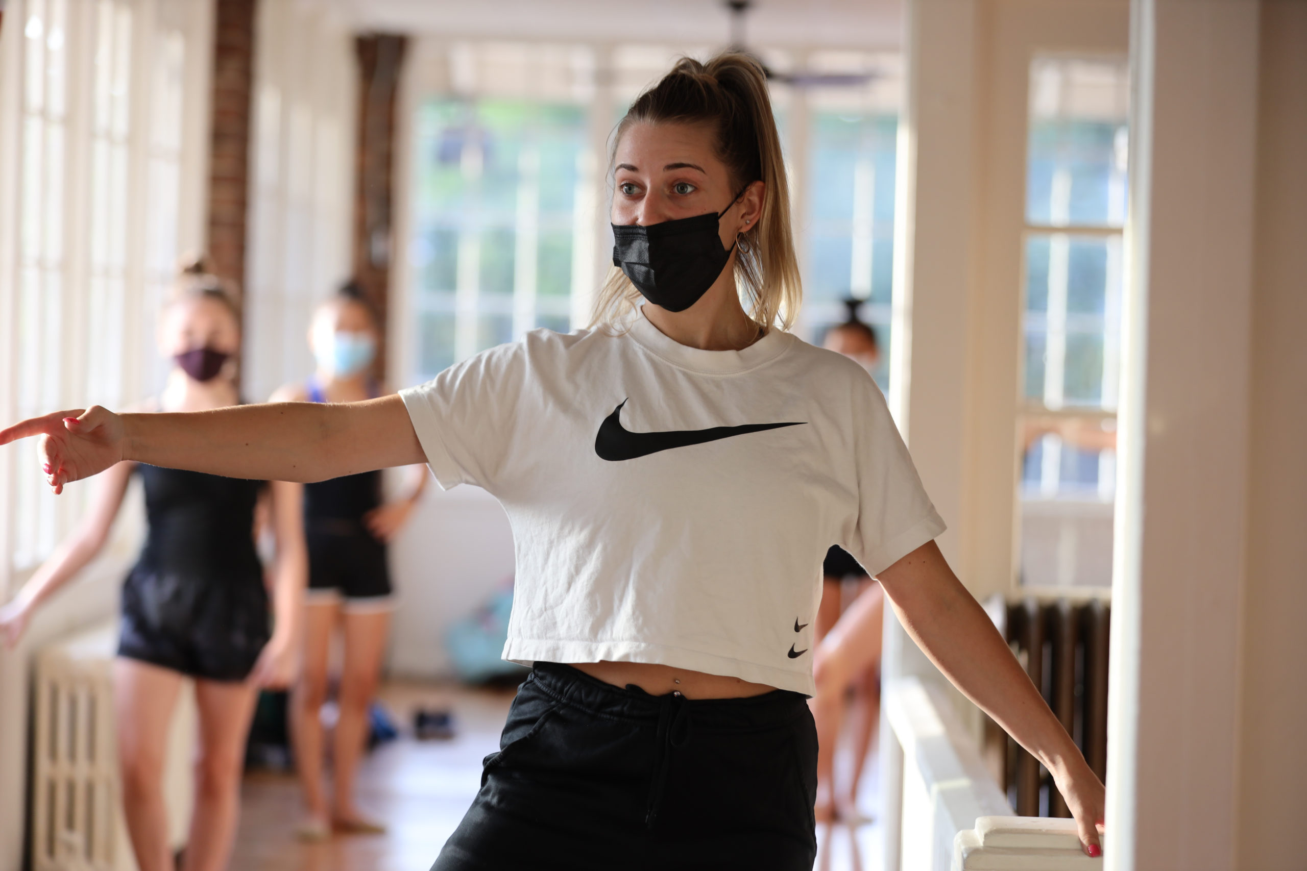 A dancer in a Nike t-shirt, sweats, and a black mask over her nose and mouth demonstrates for a group of younger dancers, one hand resting on a barre and the other in second position.