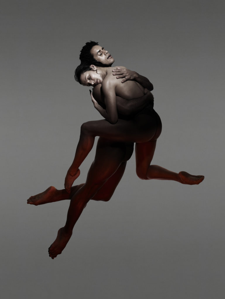 Marcelino Sambé and Francesca Hayward embrace in the air, eyes closed, wrapping their arms around the other's chest.  Their legs and feet have a beautiful classic shape.  Their bare feet and minimalist costumes give the impression of nudity.