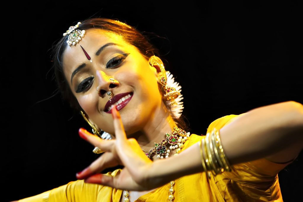A portrait of Mythili Prakash in performance. Her head is tilted and eyes downcast as she gazes, smiling, at her hand, raised to sternum height and forming a mudra with red-painted fingertips.