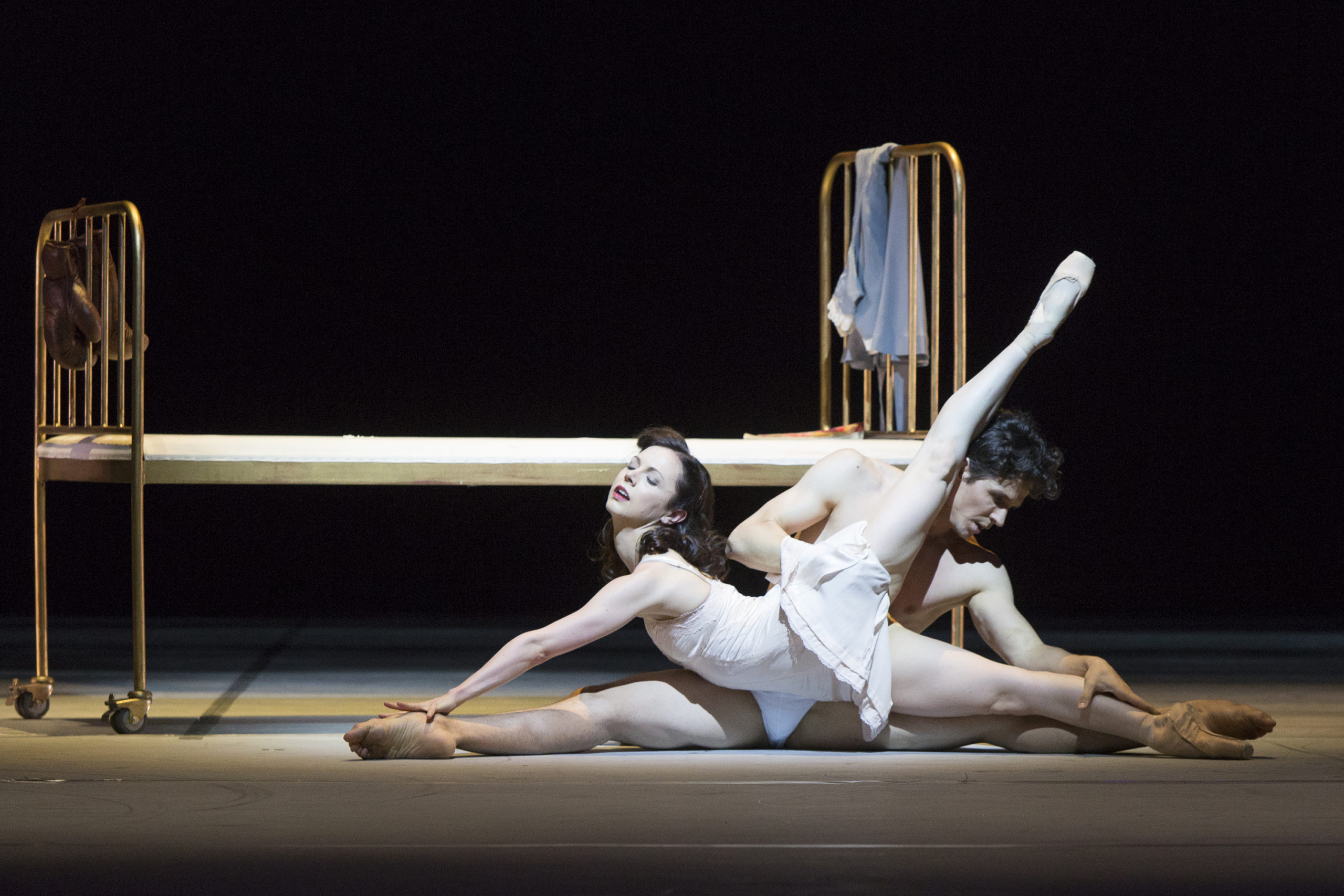 Jillian Vanstone and Guillaume Coté are white ballet dancers with dark hair. They wear delicate white costumes and lie on the stage floor in front of a small bed, facing opposite directions. Vanstone's back is arched and her left leg points back and upward.