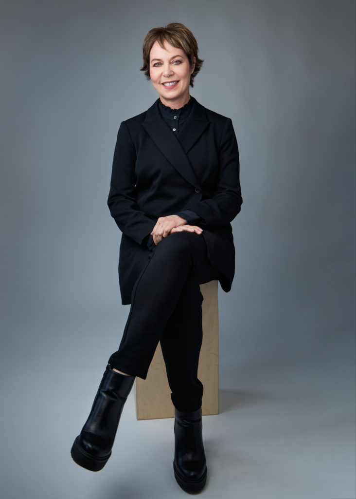 Susan Jaffe has short, layered dark hair and fair skin. She wears a black suit and sits cross-legged on a stool, facing the camera.