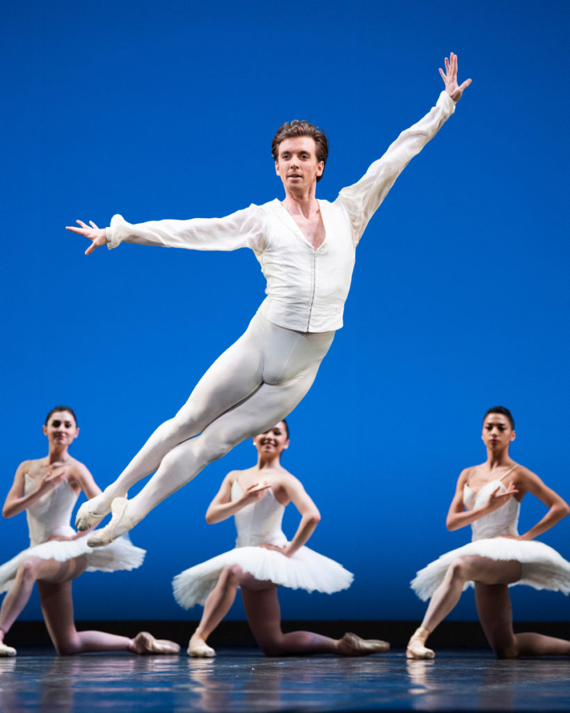 In a white long-sleeved tunic and matching tights, Ulrik Birkkjaer is captured mid-air in a cabriole back. His palms are flexed, but his gaze serene as he looks out at the audience. Behind him, a trio of dancers in simple white tutus pose, kneeling on one knee.