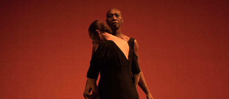 Germaine Acogny stands upright, facing downstage with her arms lightly raised from her sides. Malou Airaudo faces Acogny, chin lowered to one of her shoulders, arms wrapping around Acogny's back. The stage behind them is lit a deep red.