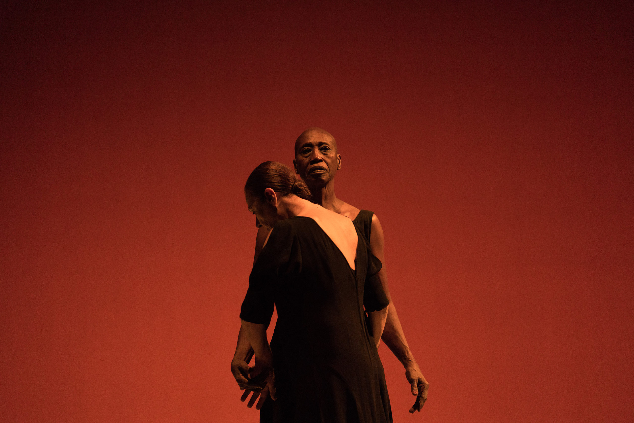 Germaine Acogny stands upright, facing downstage with her arms lightly raised from her sides. Malou Airaudo faces Acogny, chin lowered to one of her shoulders, arms wrapping around Acogny's back. The stage behind them is lit a deep red.