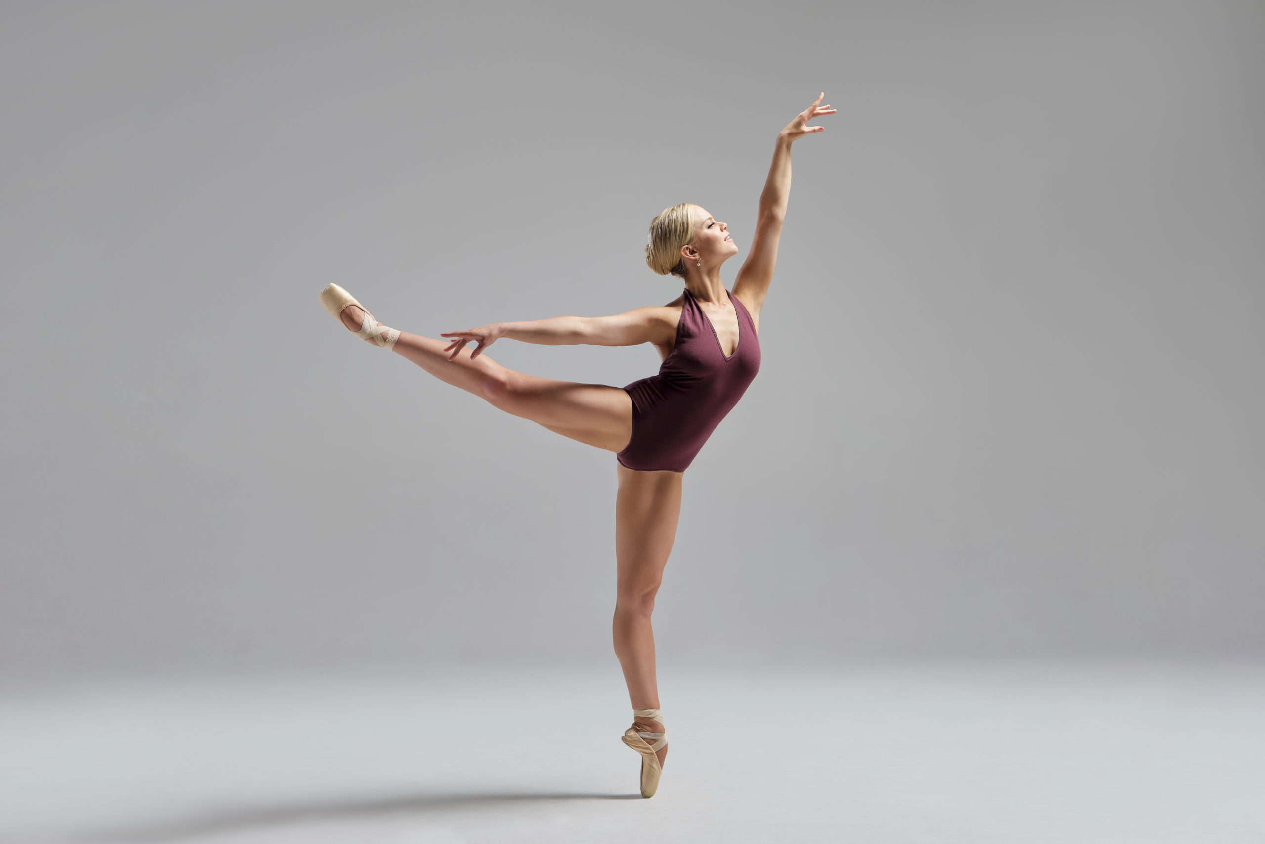 Haley Hilton is a fair-skinned woman with blonde hair pulled back into a bun and wearing a maroon sleeveless leotard. She faces left and poses in arabesque on pointe on her left leg, with her right leg and arm pointing backward and her left arm raised overhead.