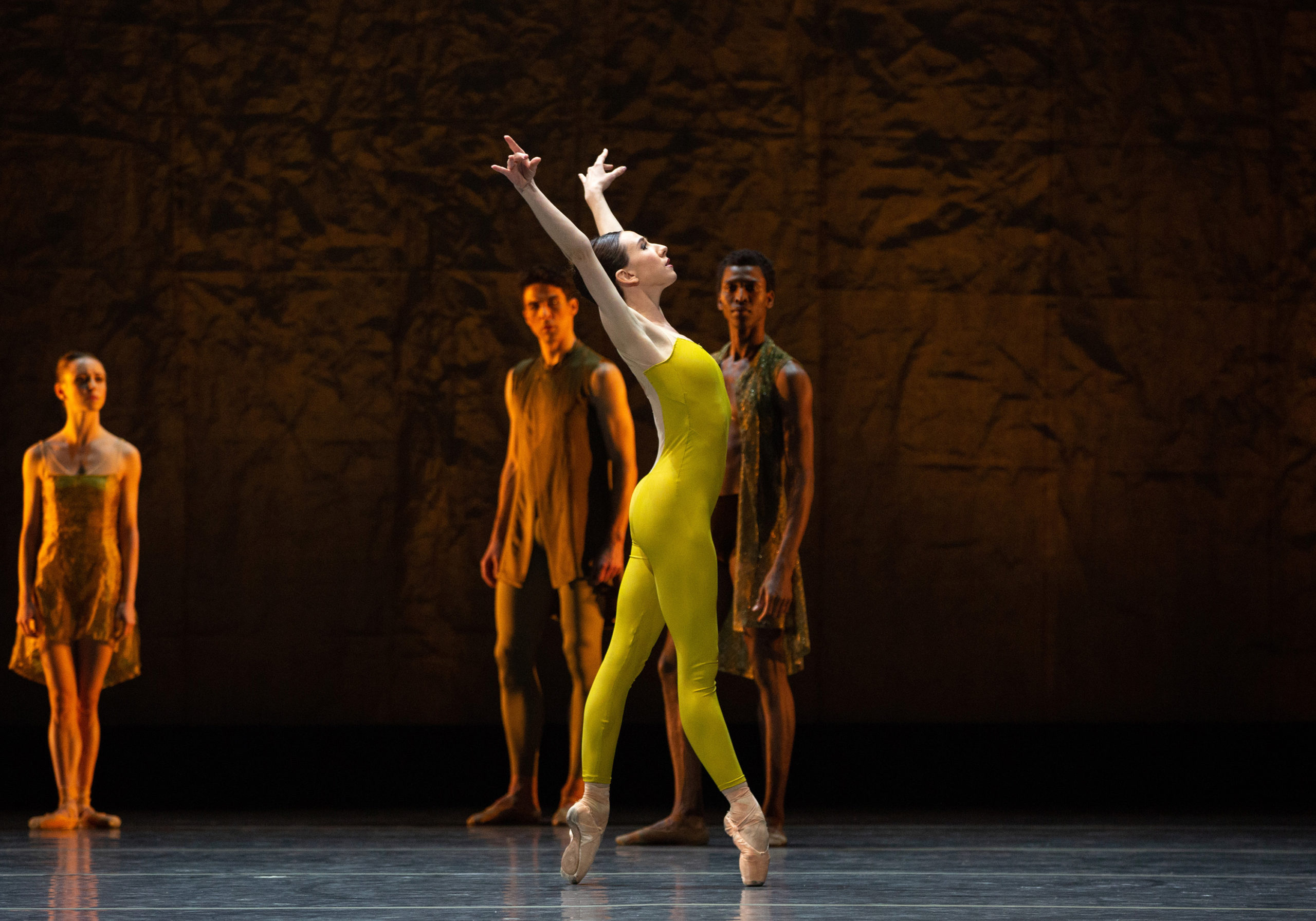 Isabella Boylston, wearing a chartreuse unitard and pointe shoes, is shown balancing in fourth position facing the side, ribs splayed as her arms rise in a V overhead, gaze lifted.