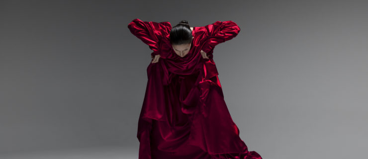 Vangeline is a female dancer with slicked-back dark hair and pale-white makeup and wearing a billowing ruby red gown, bending forward at the waist, arms bent to the sides.