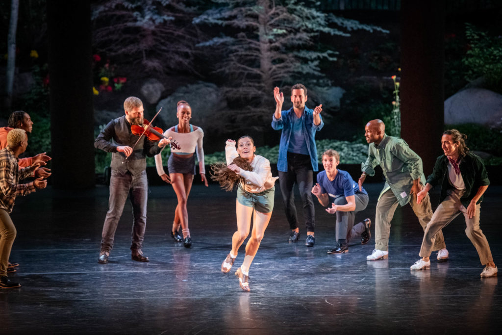 Melissa Toogood leans forward, hair and arms swinging as she taps at the center of a nature-backed stage. Circling around her are seven other dancers in tap shoes, smiling and clapping, as well as a violinist watching as he plays.