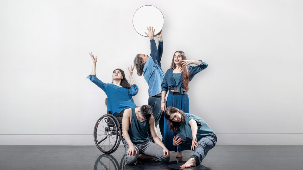 Five dancers dressed in blue pose in front of a white background.  One is on his knees, looking down, while behind him another smiles exaggeratedly broadly, gazing into the distance.  A dancer in a wheelchair gestures as if supporting something invisible above her head, while another right behind her raises a circle above her head.
