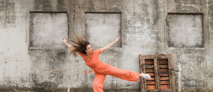 Alexandra Bodnarchuk is a white woman with long brown hair. She wears a short-sleeved orange jumpsuit with a tie waist, and she lunges forward on her bent right leg while her left leg, arms and hair extend outward.