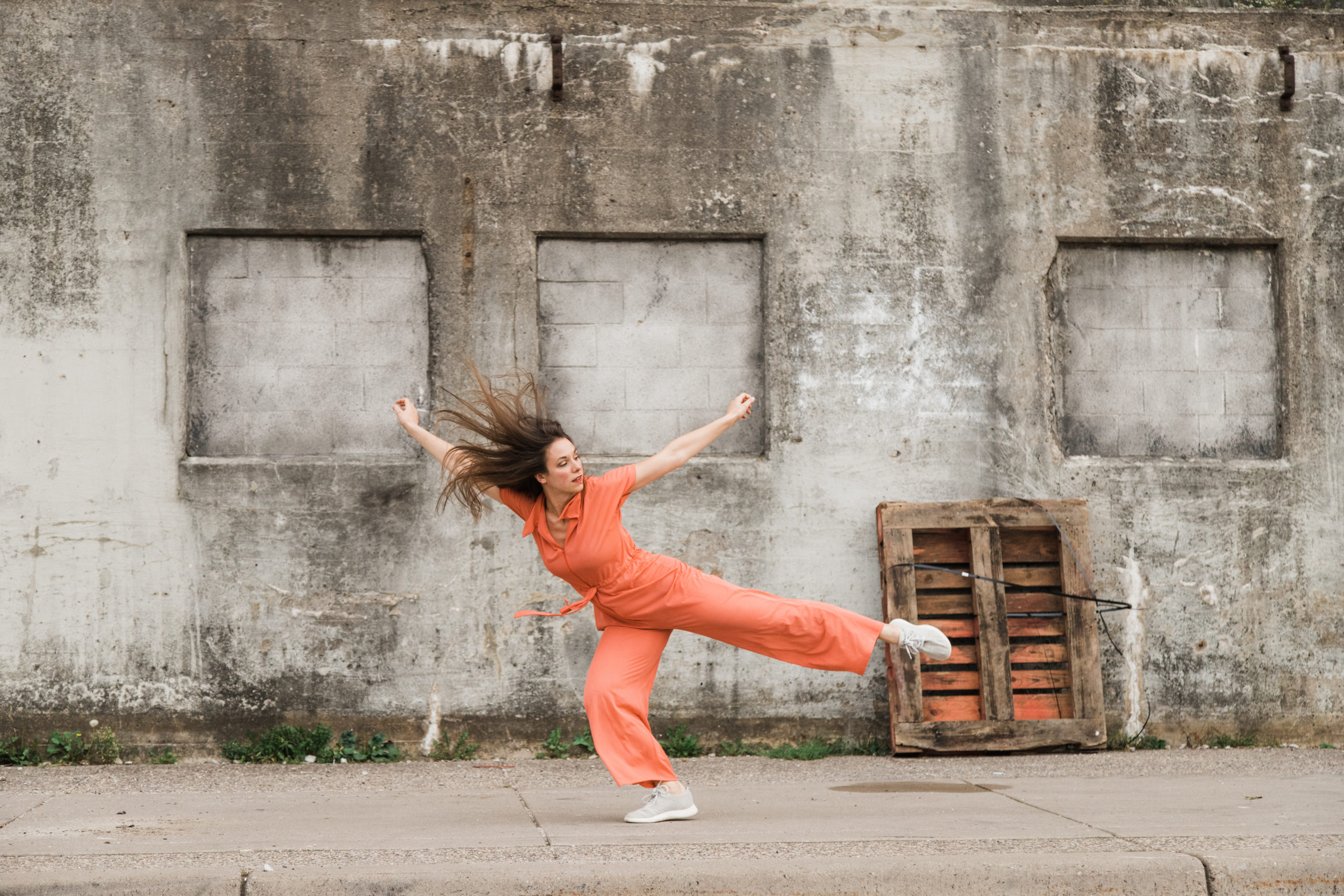 Alexandra Bodnarchuk is a white woman with long brown hair. She wears a short-sleeved orange jumpsuit with a tie waist, and she lunges forward on her bent right leg while her left leg, arms and hair extend outward.