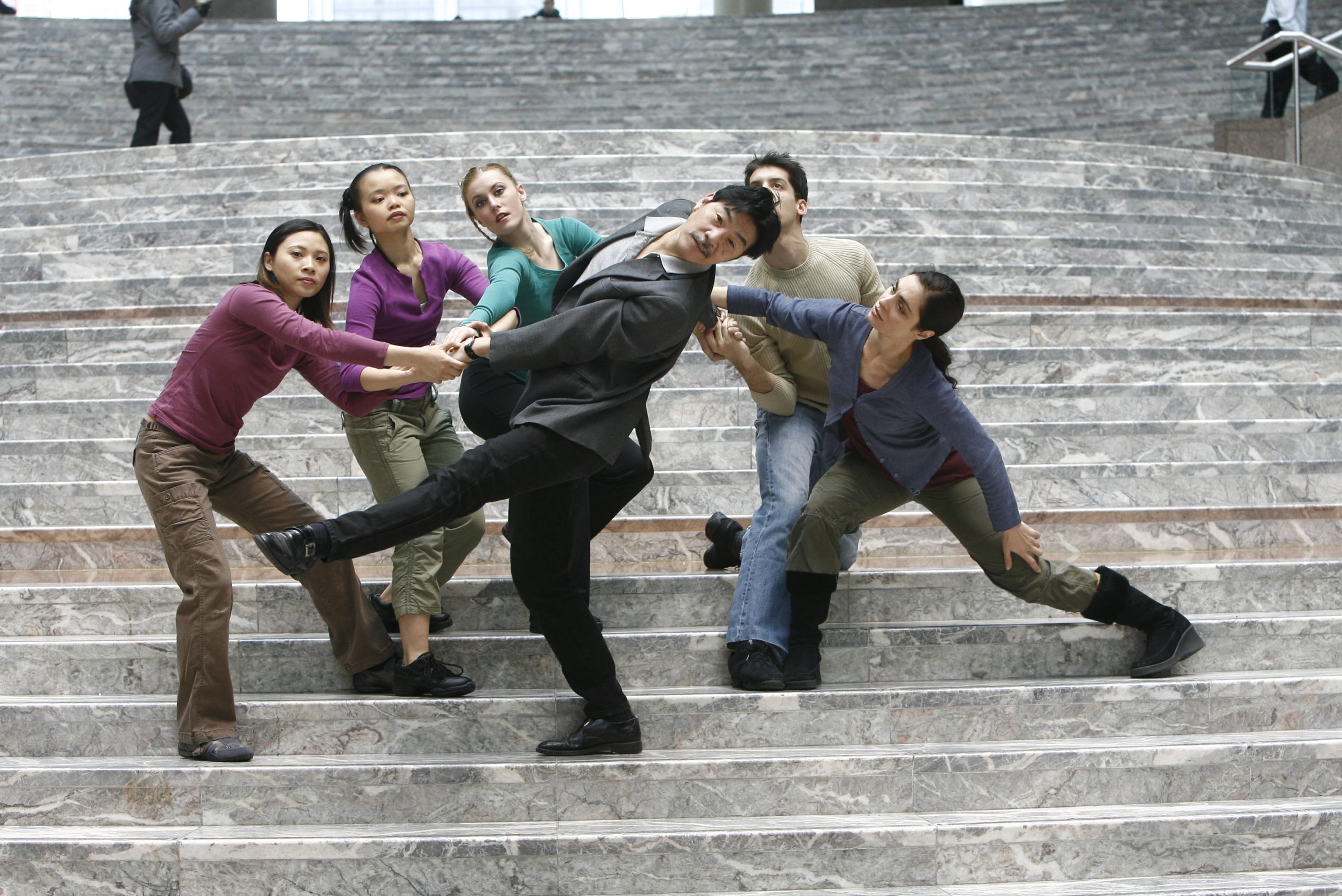 HT Chen is a middle-aged Asian man wearing a gray blazer and black pants. He leans back and is supported by five young dancers wearing muted colors, on the cement steps of the Winter Garden in New York City.