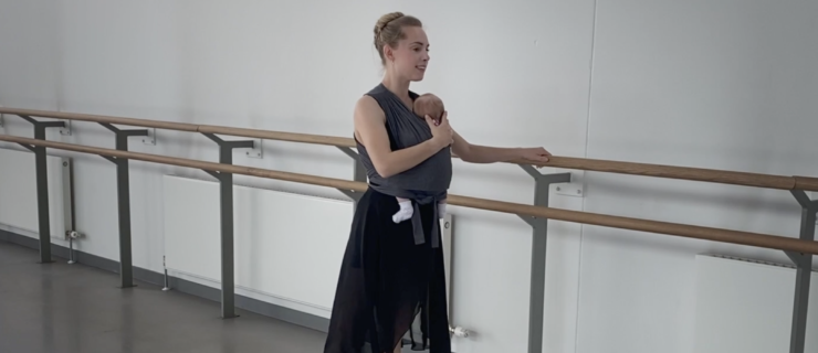 Bethany Kingsley-Garner stands in tendu second, her left hand on the barre and her right hand cradling the head of her newborn, who is swaddled in a carrier strapped to her chest.