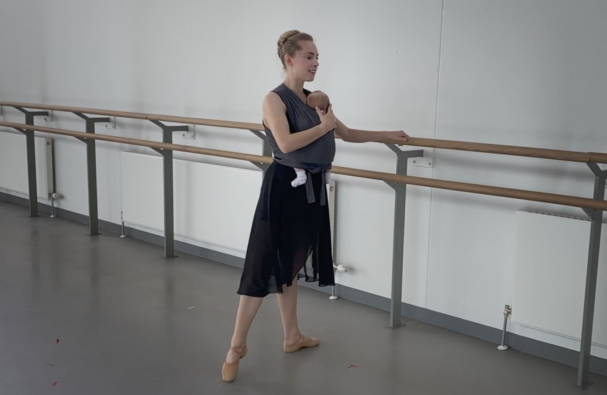 Bethany Kingsley-Garner stands in tendu second, her left hand on the barre and her right hand cradling the head of her newborn, who is swaddled in a carrier strapped to her chest.