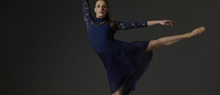 Courtney Nitting is a fair-skinned woman with long red hair pulled into a bun. She wears a long-sleeved blue dress that falls just above the knee and does an arabesque toward the right with her left leg pointing back.