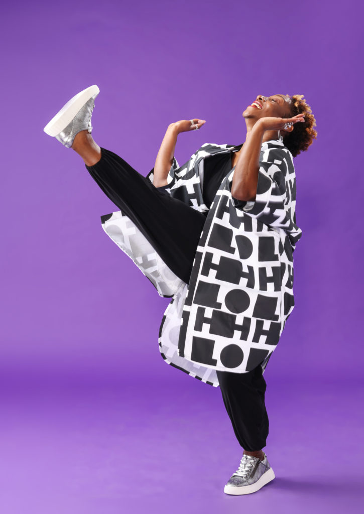 Against a bright purple backdrop, LaTasha Barnes smiles widely, eyes shut as she tips her head back to the ceiling. One leg is extended high in front of her with a flexed foot. Her arms bend at the elbows in front of her ribs, palms upraised as though lifting an invisible string tied to her foot. She wears a black jumpsuit, black and white patterned jacket, and silver sneakers.