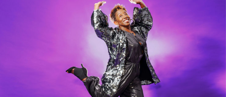 In front of a bright purple backdrop, and with a big smile, LaTasha Barnes is wearing a sparkling cardigan, metallic gray pants, and black high heels as she lifts both arms, and kicks a leg back.