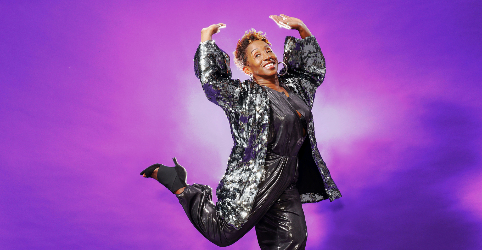 In front of a bright purple backdrop, and with a big smile, LaTasha Barnes is wearing a sparkling cardigan, metallic gray pants, and black high heels as she lifts both arms, and kicks a leg back.