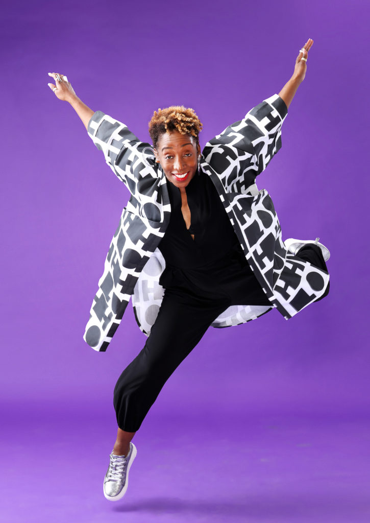 In front of a bright purple background, LaTasha Barnes smiles at the camera as she hovers just above the ground. One leg is kicked up behind her as her arms fly into a V above and behind her shoulders. She wears a black jumpsuit, black and white patterned open jacket, and silver sneakers.