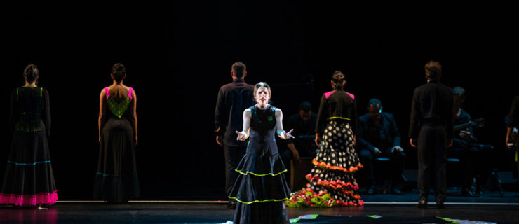 A female dancer in a long black dress stands midstage under a spotlight, facing the camera with her arms bent and fists clenched.