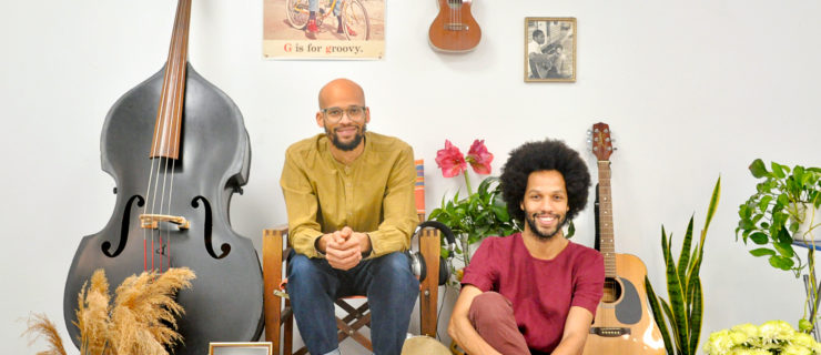 Gregory Richardson is a light-skinned Black man who is bald and has a beard and glasses. Leonardo Sandoval is a man with a medium complexion, curly hair and a beard. They sit on a carpet surround by musical instruments.