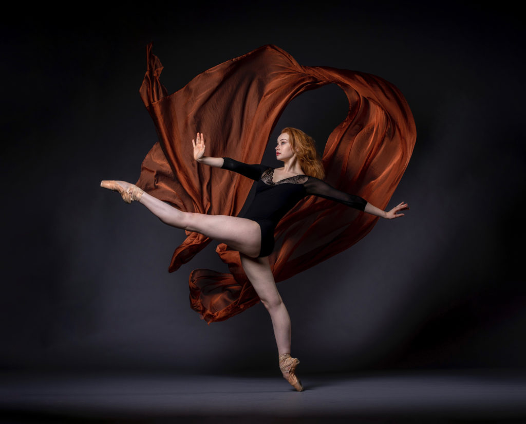 Graceanne Pierce is a fair-skinned woman with red hair. She wears pointe shoes and a black leotard. She faces left, balancing on pointe on her right leg and kicking up her left leg, framed by billowing copper fabric.